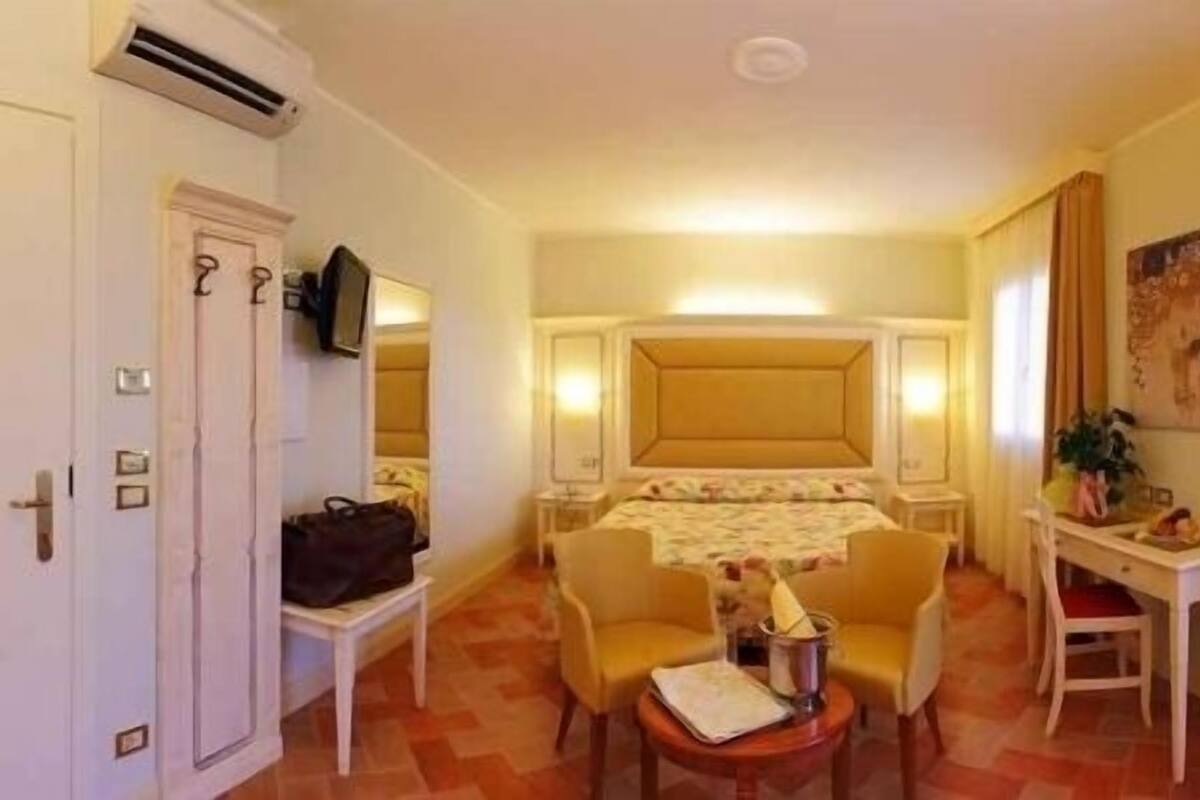 Quarto de hotel com cama, duas cadeiras, uma mesinha, escrivaninha, televisão e janela com cortinas.