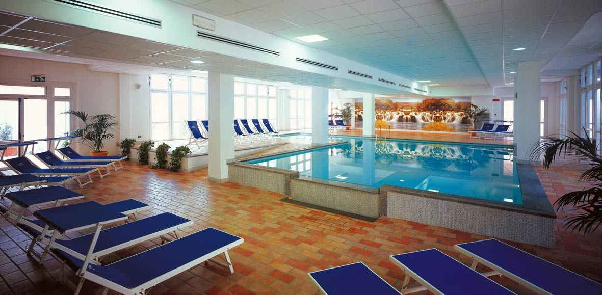 An indoor swimming pool with blue lounge chairs and a tiled floor.