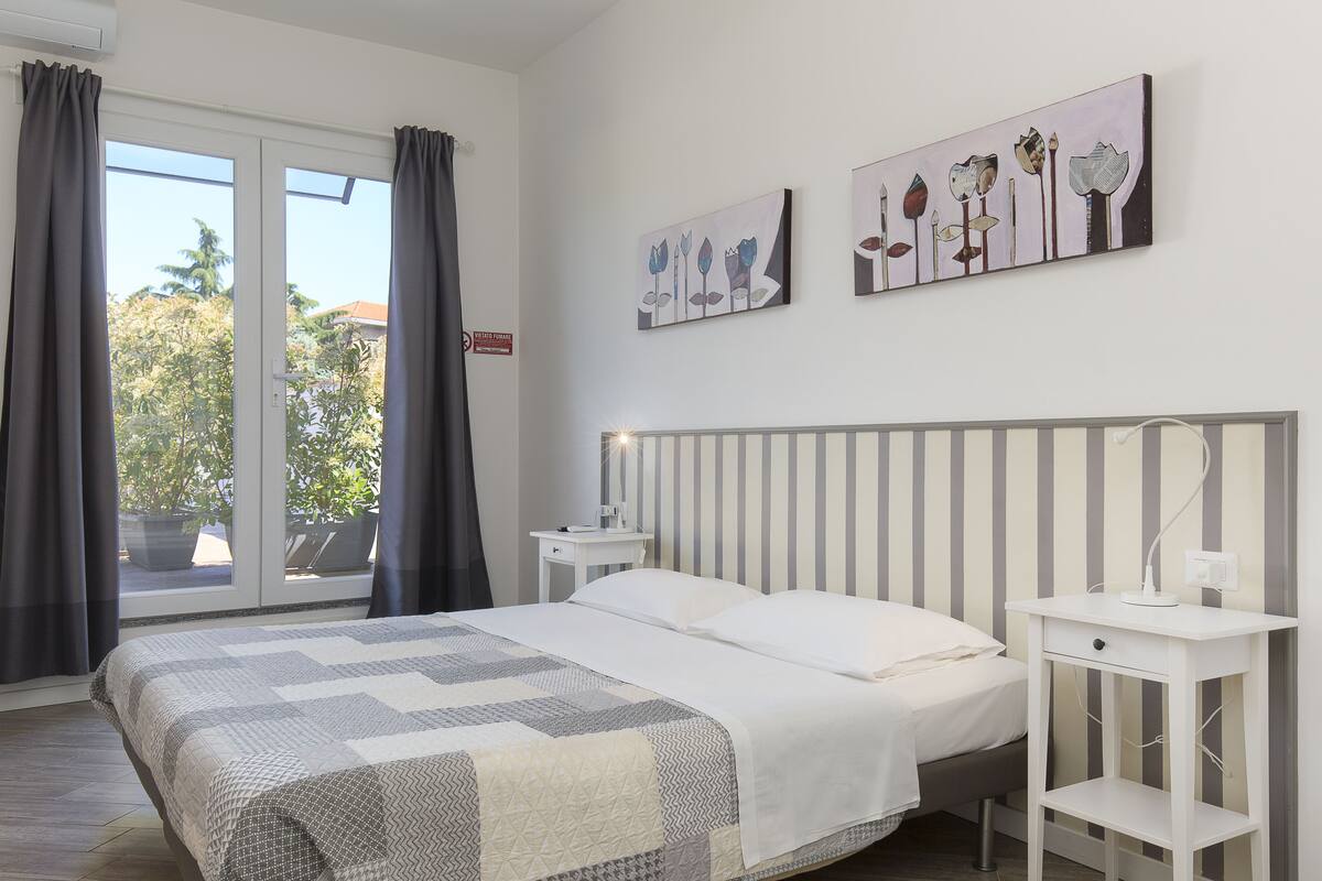 Un dormitorio con cama, mesita de noche y ventana con cortinas. Dos obras de arte decoran la pared sobre la cama.
