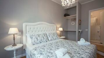 Une chambre avec un lit blanc, une literie à motifs floraux, deux lampes de chevet, un lustre, une salle de bain avec douche et une serviette sur le lit.