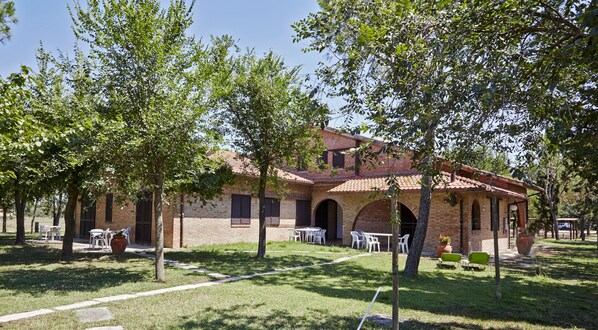 A brick building with a tiled roof, surrounded by trees and outdoor seating. - Appartamenti la Principina - I Casali (Grosseto)