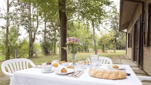 An outdoor dining table set with plates, cutlery, glasses, and a loaf of bread, surrounded by trees and a brick building.