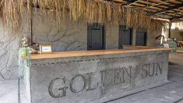 Un mostrador de recepción con el letrero 'GOLDEN SUN', fachada de piedra, una barra de madera y techo de paja.