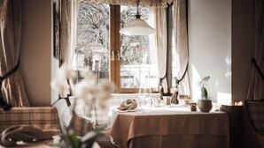 A cozy room with a dining table set for two, a window with curtains, and a hanging light fixture. - Romantik Hotel & Restaurant Stafler (Campo di Trens)