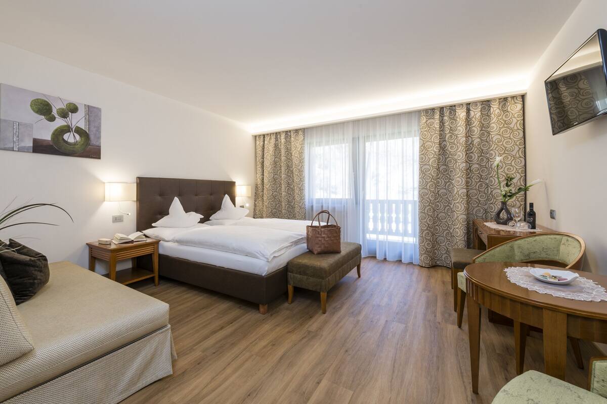 A hotel room with a bed, bedside table, lamp, bedside plant, a small table with a chair, and a dining table with a chair.