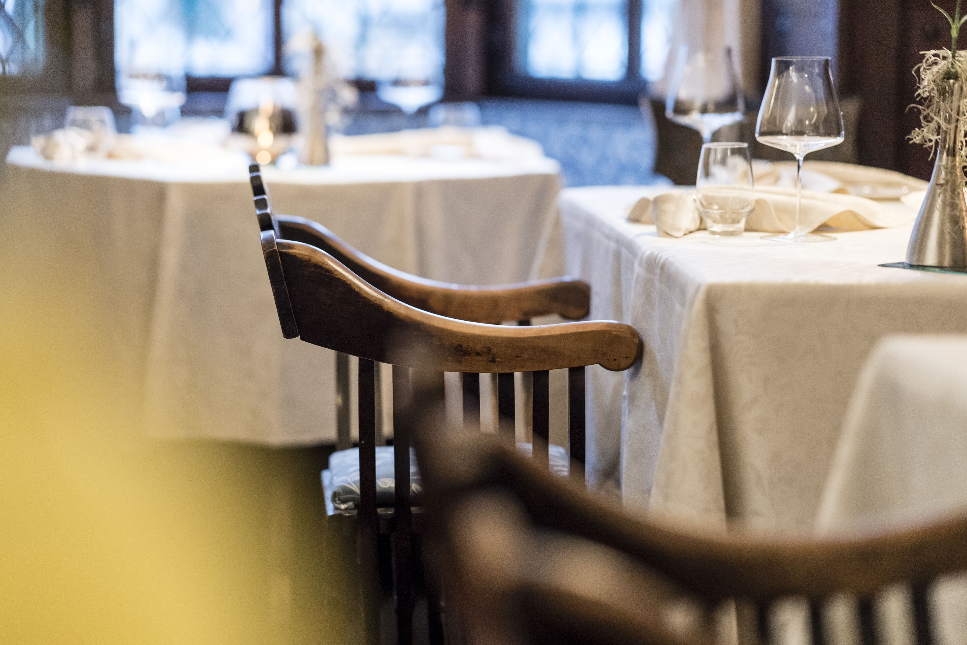 A dining area with round tables covered in white tablecloths, wooden chairs, and wine glasses.
