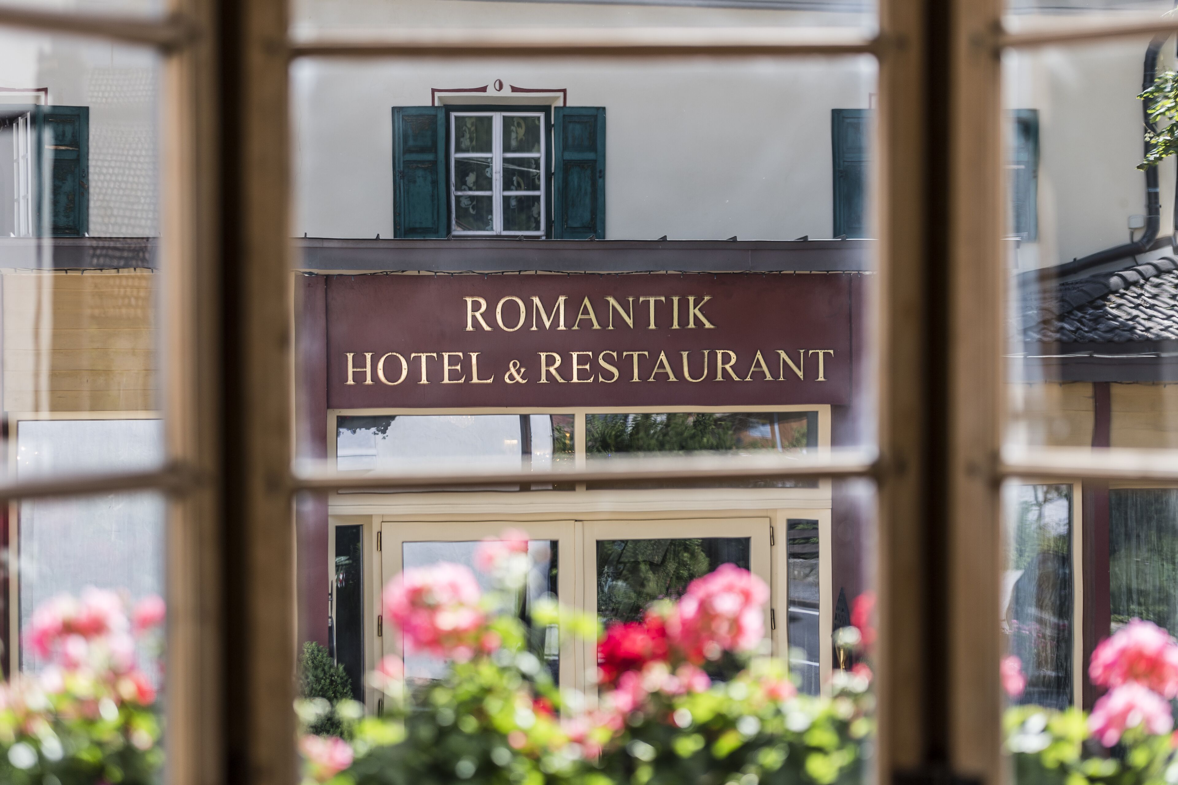A sign for 'Romantik Hotel & Restaurant' with a window view of a building and pink flowers in the foreground.