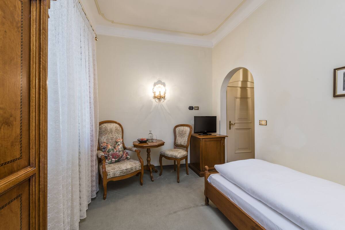 A hotel room with a bed, two chairs, a small table with a glass, a television, and a wall-mounted light fixture.