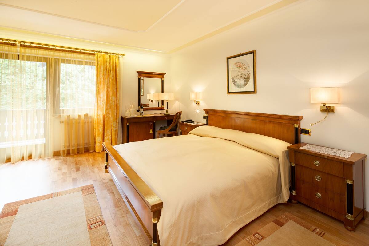 A well-lit bedroom with a large bed, a wooden nightstand, a desk with a chair, and a window with curtains.