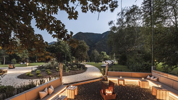 An outdoor seating area with a fire pit, surrounded by a landscaped garden and a view of a mountain range. - Romantik Hotel & Restaurant Stafler (Campo di Trens)