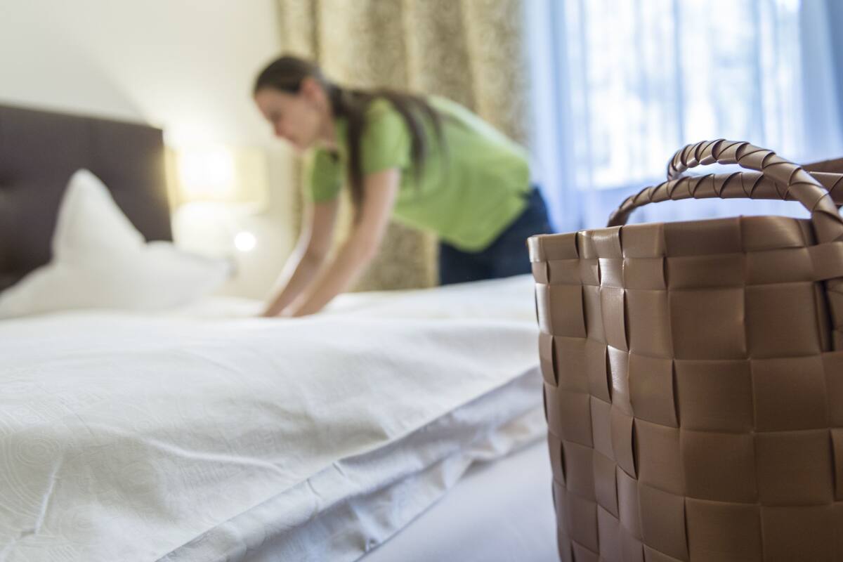 A hotel room with a neatly made bed, a large woven basket, and a person in a green shirt cleaning the bed.