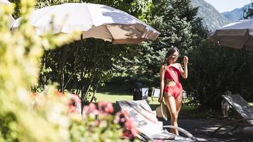 A woman in a red swimsuit standing under a white umbrella in a garden with lounge chairs and a statue.