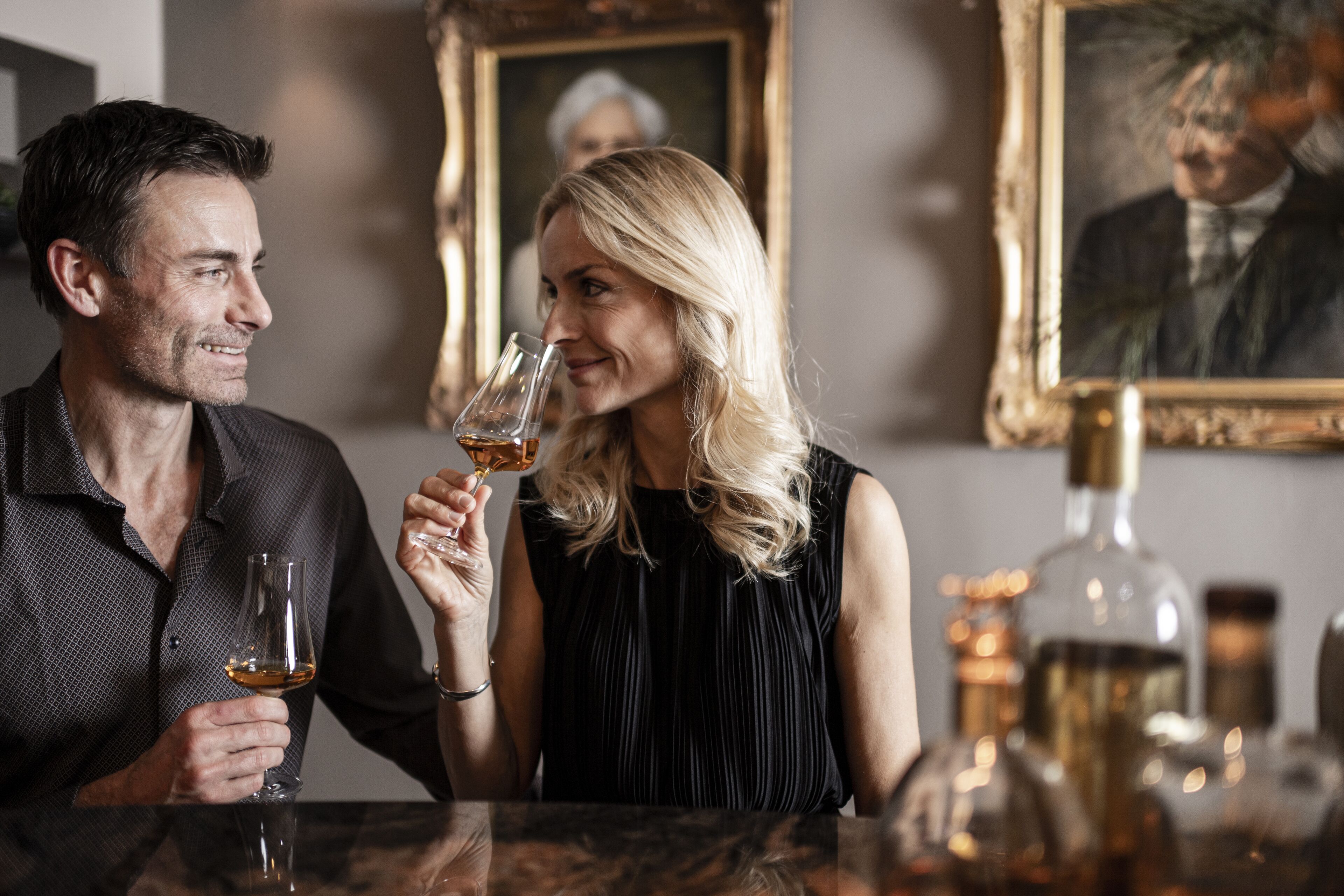 Dos personas sentadas en una mesa con una copa de vino, una botella de licor y cuadros en la pared.