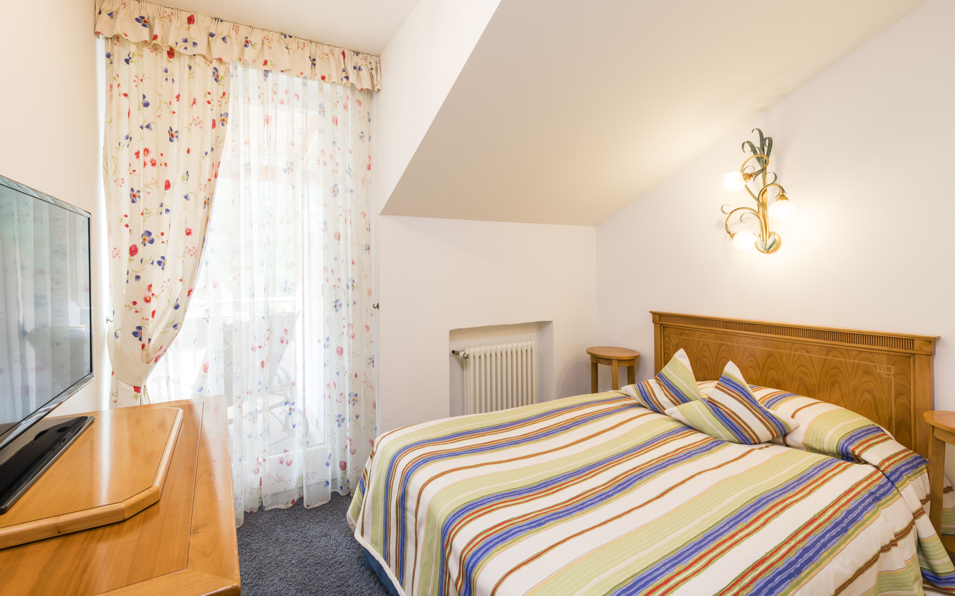 Une chambre d’hôtel avec un lit, un bureau, un radiateur et un luminaire fixé au mur.