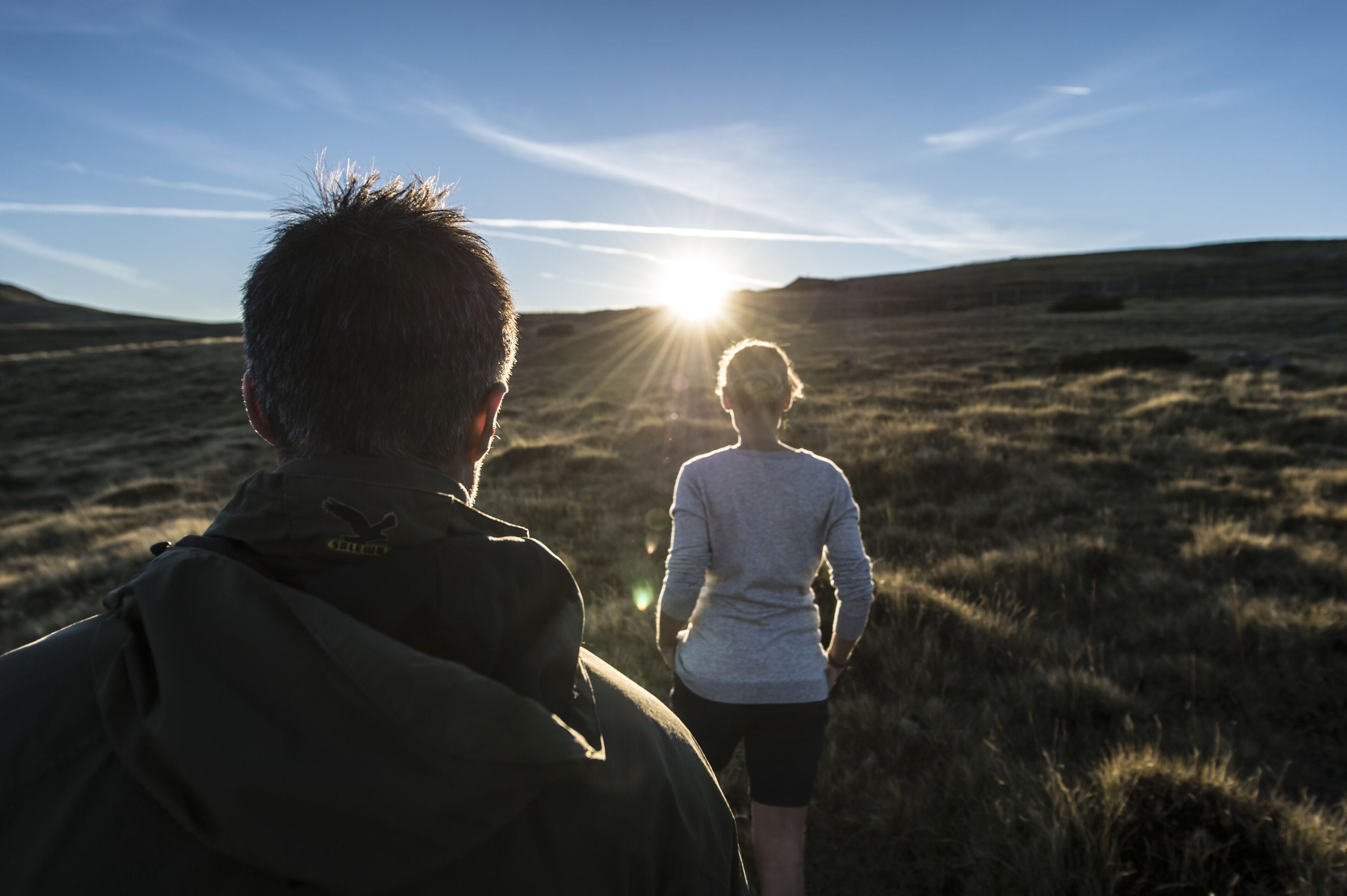 A person in a dark jacket walking towards the sun with another person in a light-colored top and dark pants.