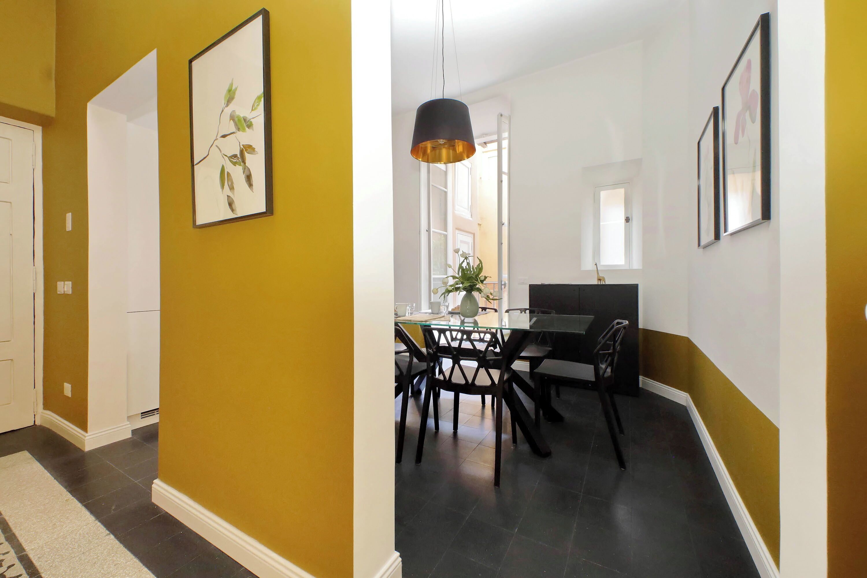 A hallway with a yellow wall, a dining area with a glass table and chairs, and framed artwork on the walls.
