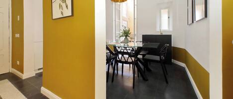 A hallway with a yellow wall, a dining area with a glass table and chairs, and framed artwork on the walls.