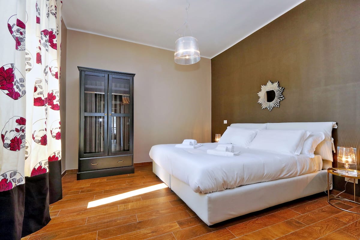 A bedroom with a large bed, a wardrobe, a hanging light fixture, and a wall-mounted mirror.