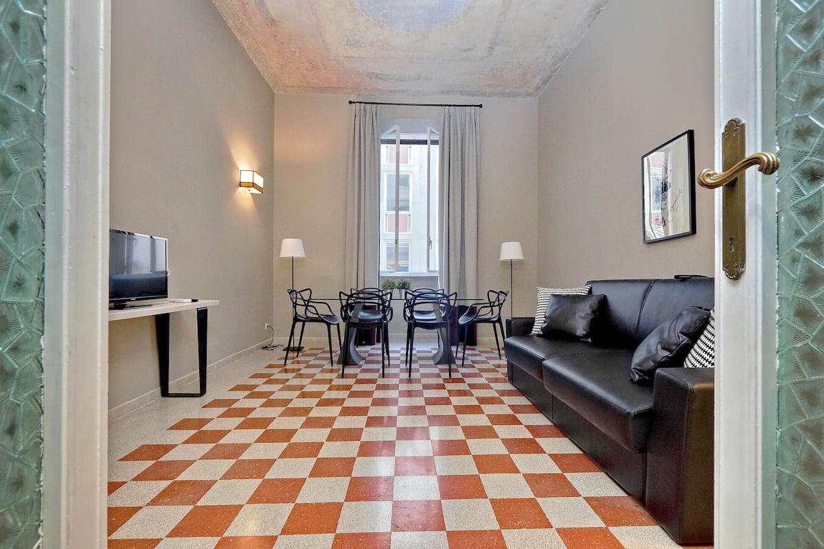 A room with a checkered floor, a black leather sofa, a TV on a stand, a dining table with chairs, and a window with curtains.