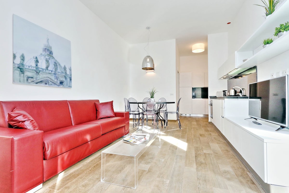 A modern living room with a red sofa, a glass coffee table, and a dining area with a chandelier.