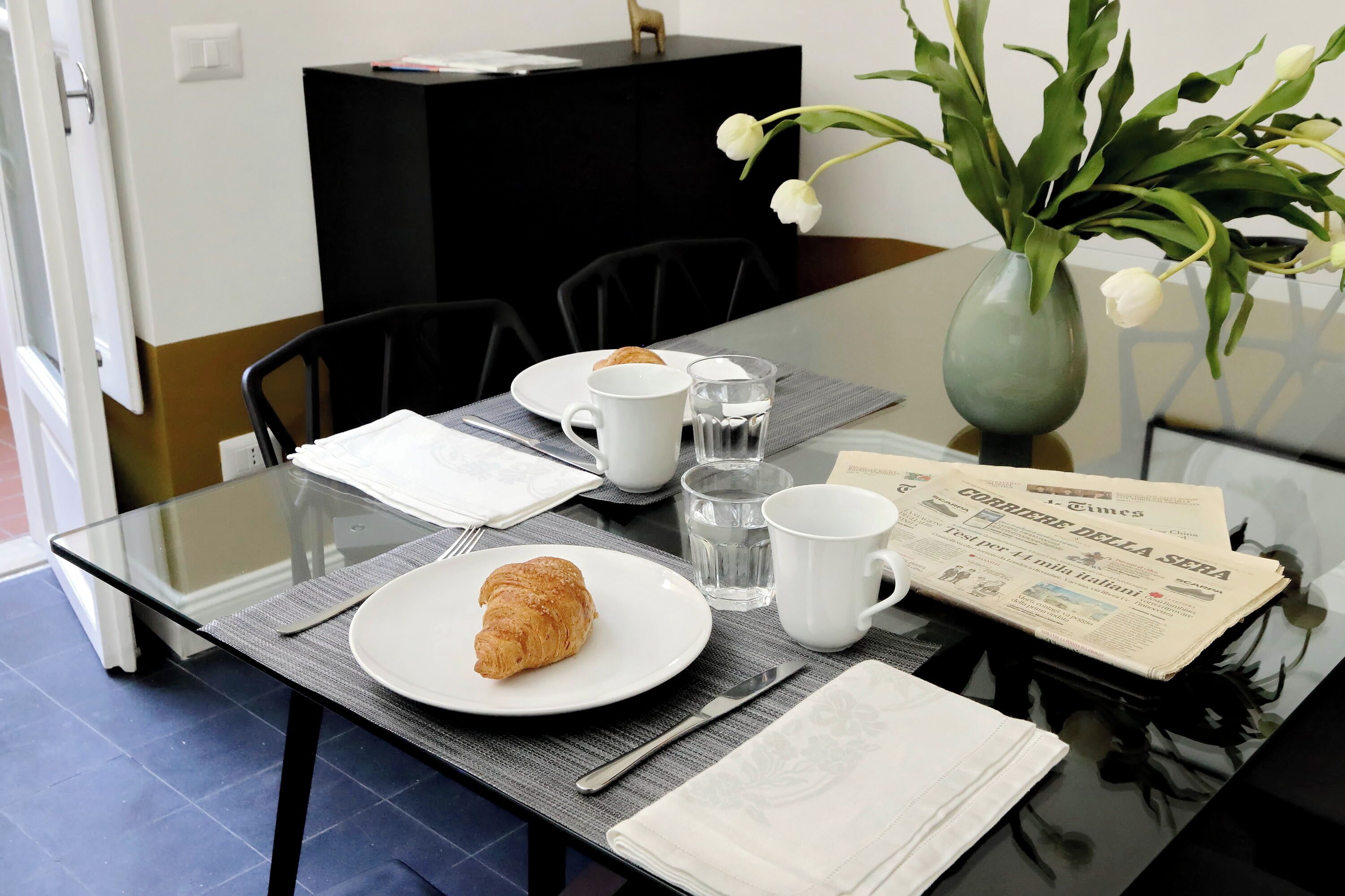 Una mesa de comedor con un croissant, un periódico y un florero con flores.