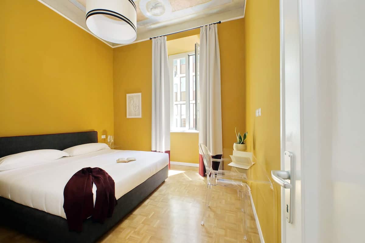 A hotel room with a bed, a bedside table, a chair, a window with curtains, and a view of the outside.