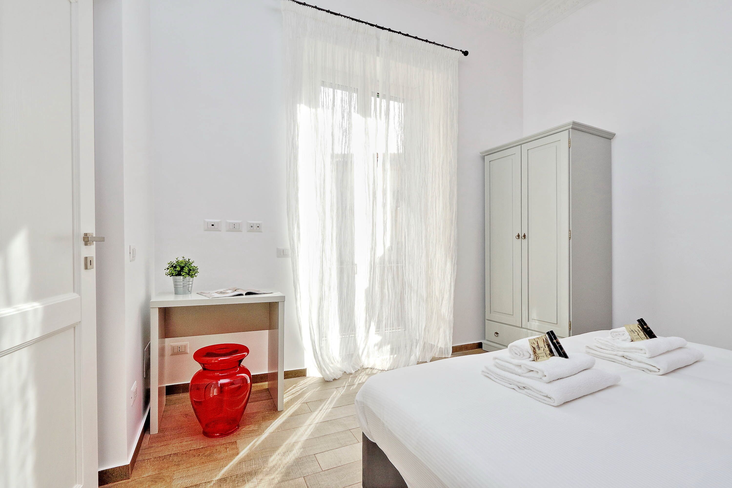 Ein Hotelzimmer mit einem Bett, einem Schreibtisch, einer roten Vase, einem weißen Schrank und durchsichtigen Vorhängen.