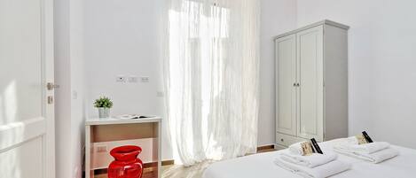 A hotel room with a bed, a desk, a red vase, a white cabinet, and sheer curtains.