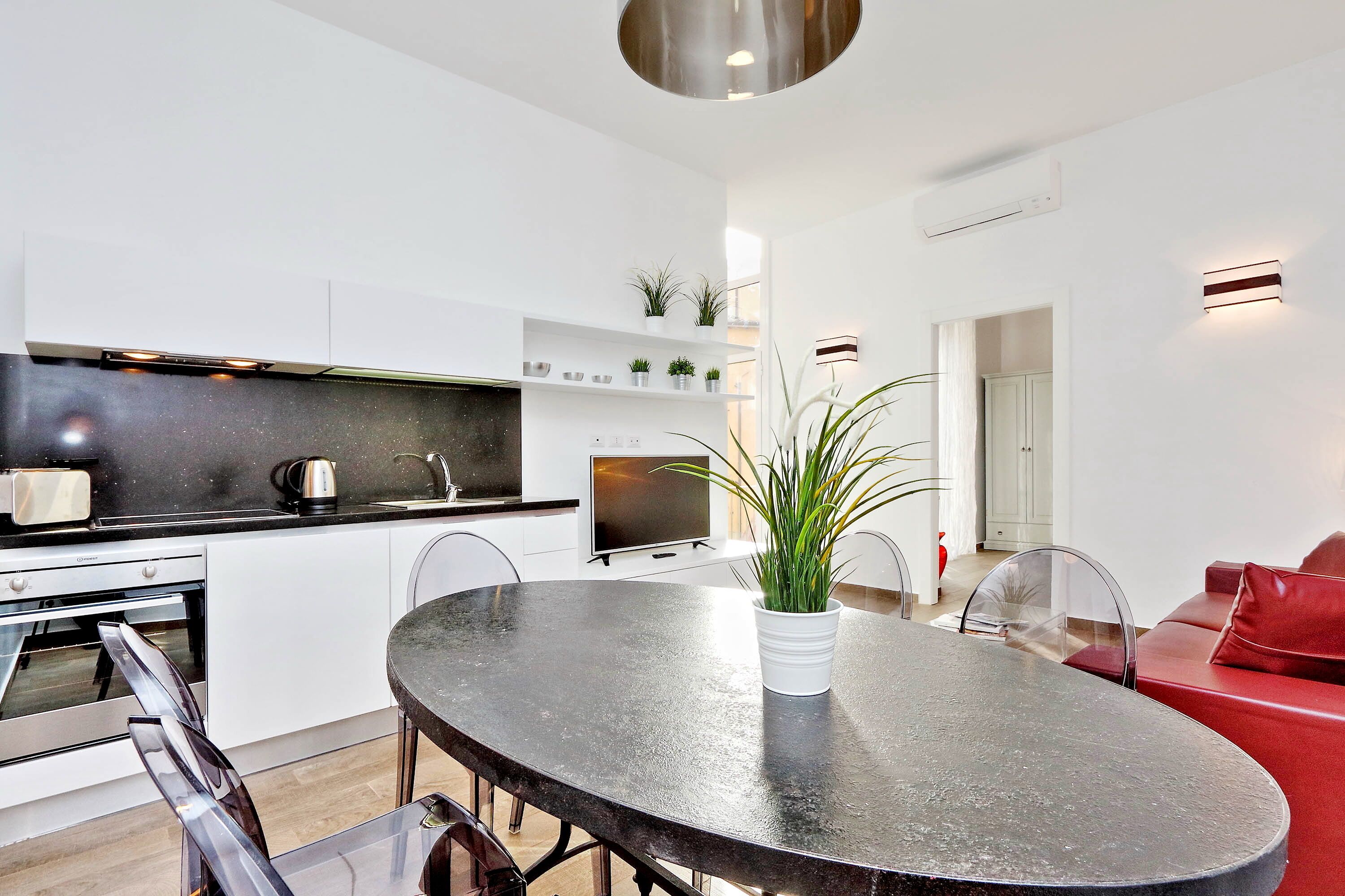 A modern kitchen with white cabinetry, a black countertop, and a built-in oven. There is a dining area with a round table and chairs, and a red sofa in the background.