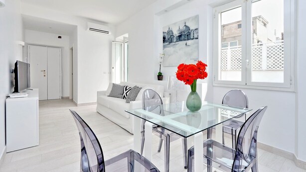 A modern living room with a glass dining table, white sofas, and a large window with a view of buildings. - Monti Apartments - My Extra Home (Rome)