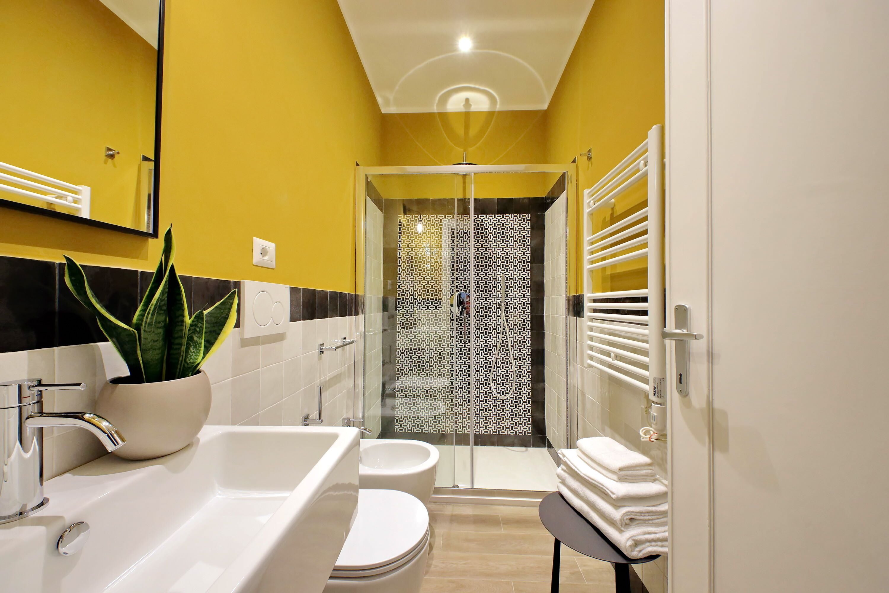 A bathroom with a yellow wall, a white sink, a toilet, a glass shower enclosure, and a towel rack.