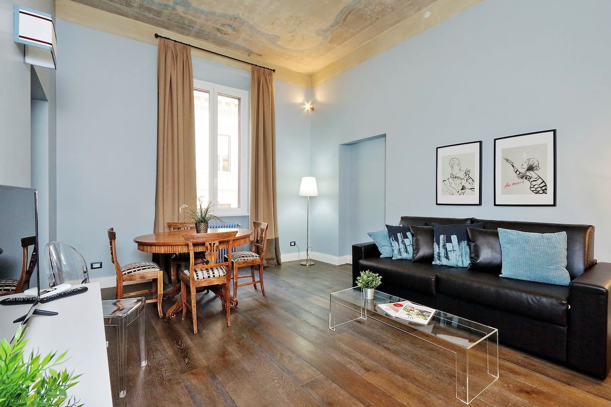 A living room with a wooden dining table, chairs, a black leather sofa, and a clear coffee table.