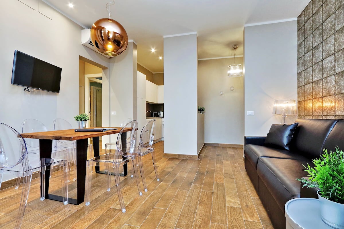 A modern living room with a wooden dining table, clear chairs, a black leather sofa, and a mounted TV.