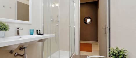 A modern bathroom with a glass shower enclosure, a white sink, and a small seating area with a cushion and a potted plant.