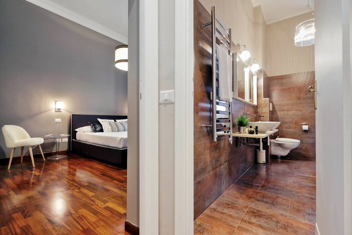A modern bedroom with a bed, bedside table, and a chair. Adjacent to the bedroom is a bathroom with a sink, mirror, and toilet.