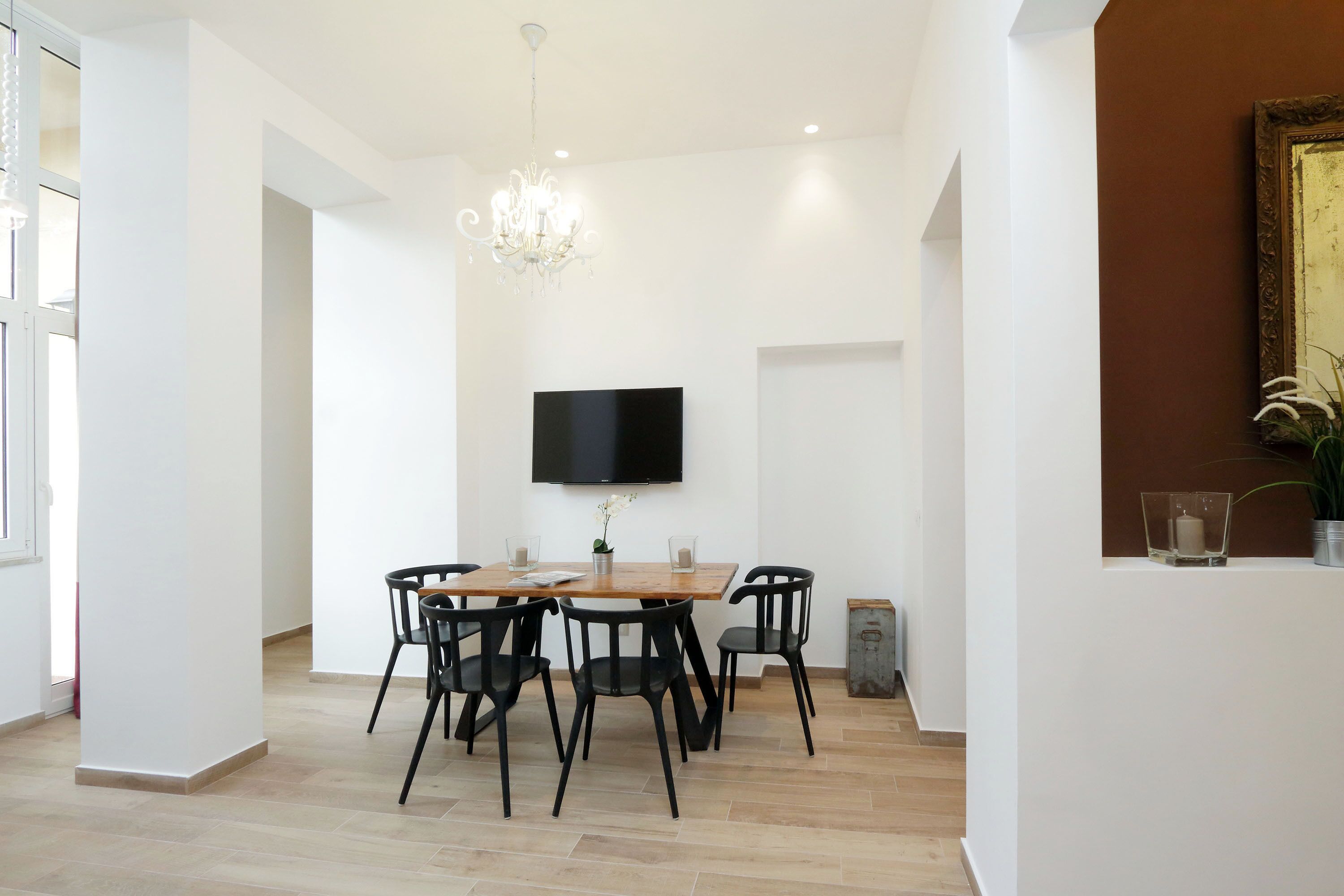 A modern dining area with a wooden table, black chairs, a chandelier, and a flat-screen TV mounted on the wall.