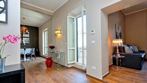 A spacious living area with a wooden floor, a glass coffee table, a red vase, and a black sofa with patterned cushions. - Monti Apartments - My Extra Home (Rome)