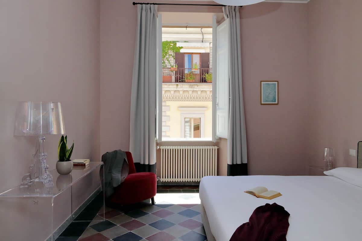 A hotel room with a bed, a red chair, a clear desk, a potted plant, and a window with a view of a balcony.