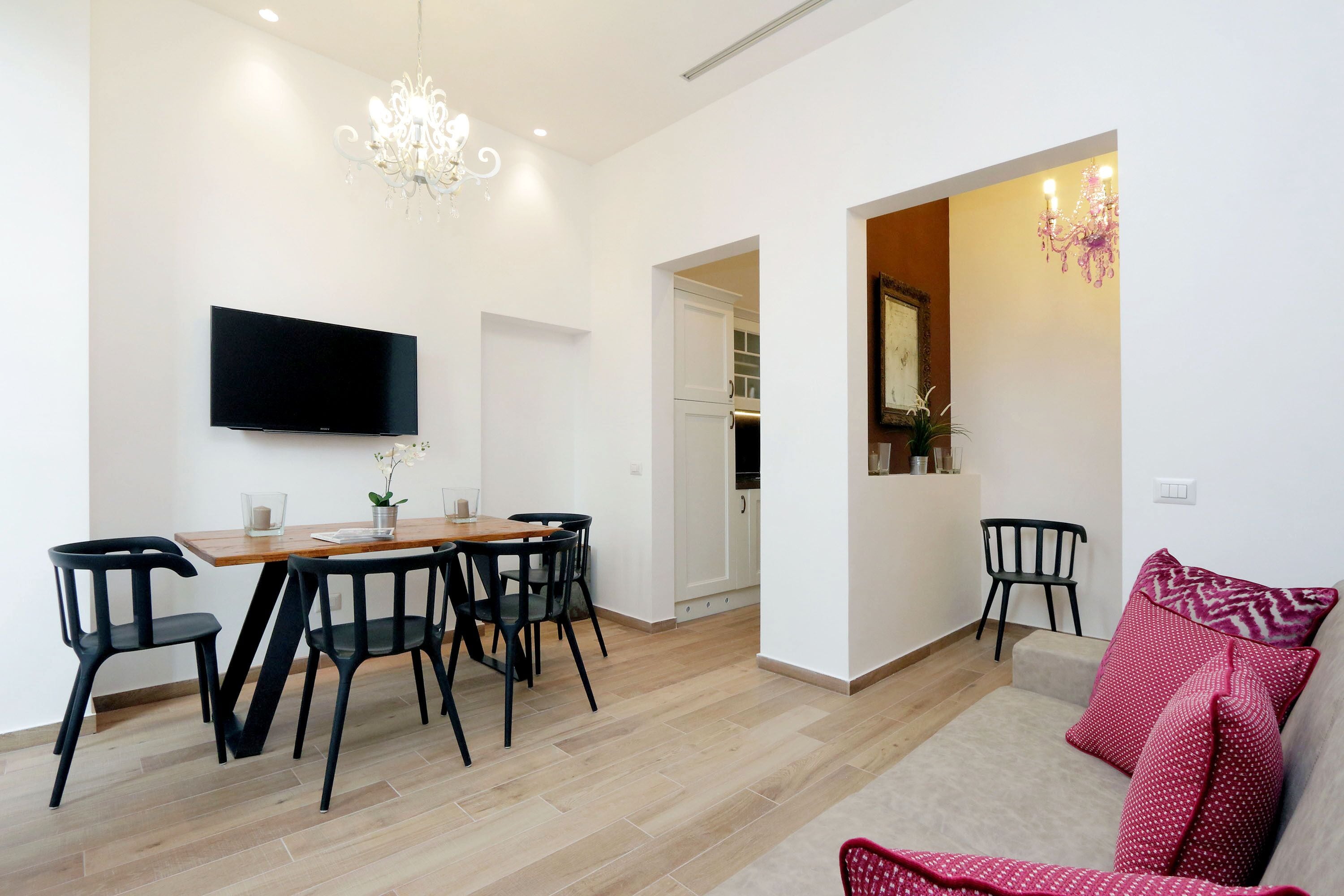 A modern living room with a dining area, a flat-screen TV mounted on the wall, and a chandelier.