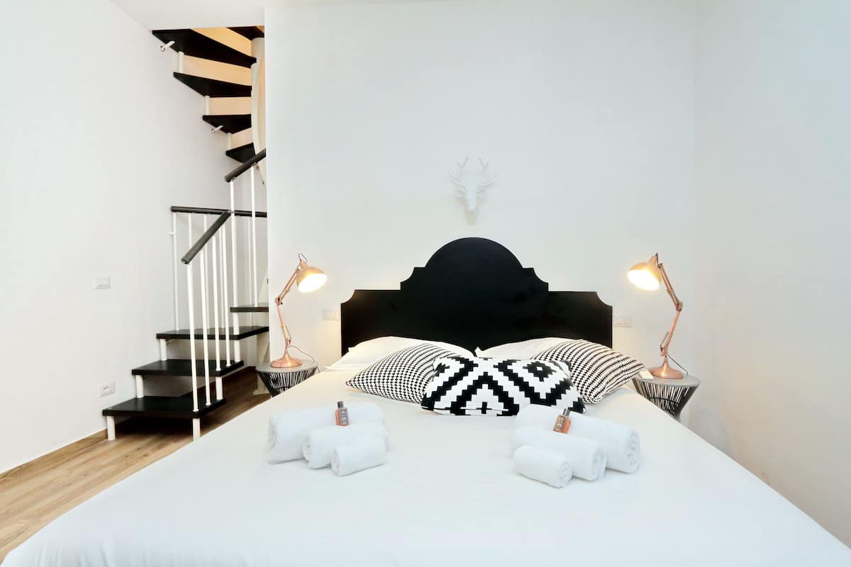 A bedroom with a bed, two bedside lamps, a headboard, a wall-mounted deer head decoration, a staircase with black and white steps, and two towels on the bed.