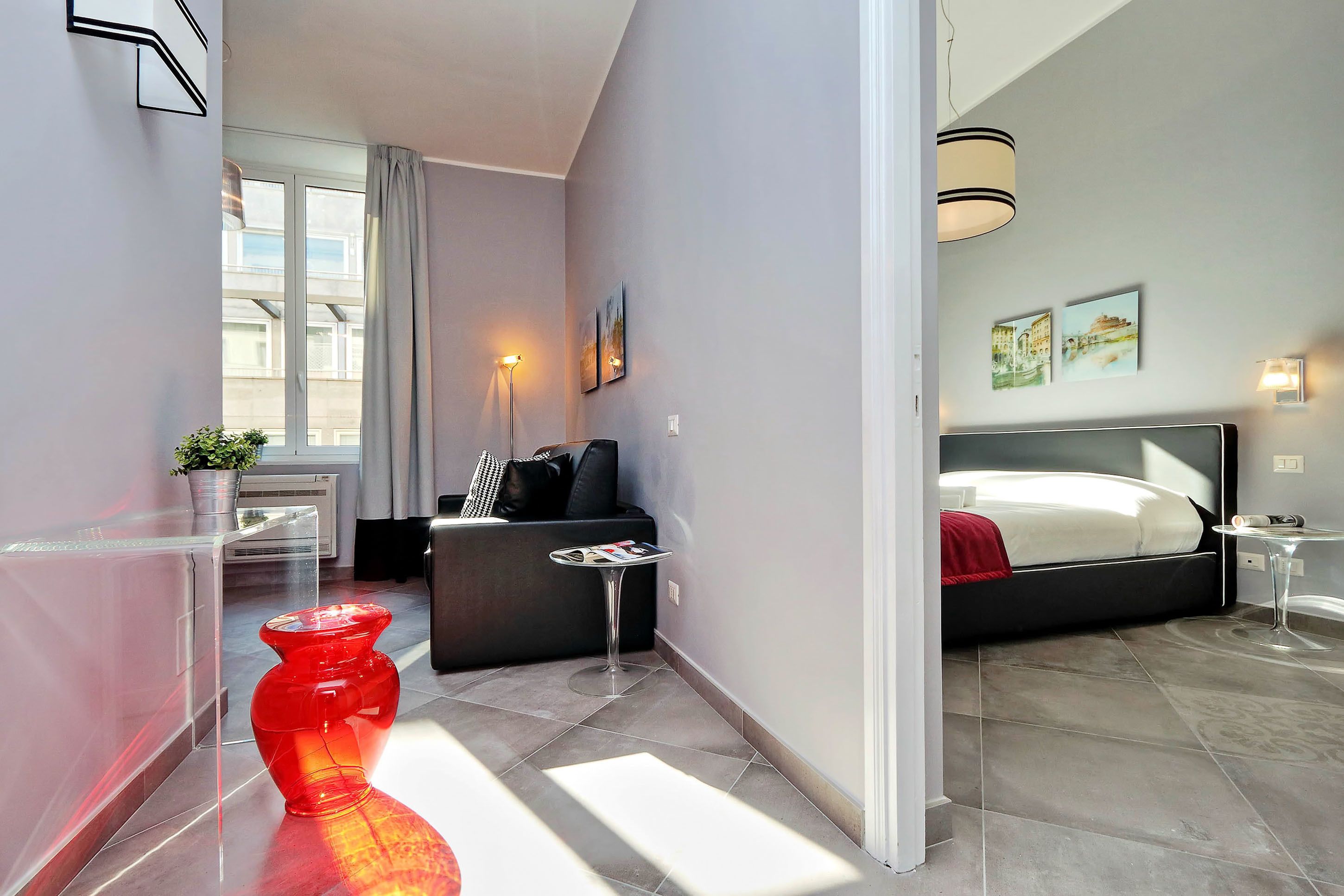 A modern hotel room with a bed, bedside table, lamp, and a red vase on the floor.
