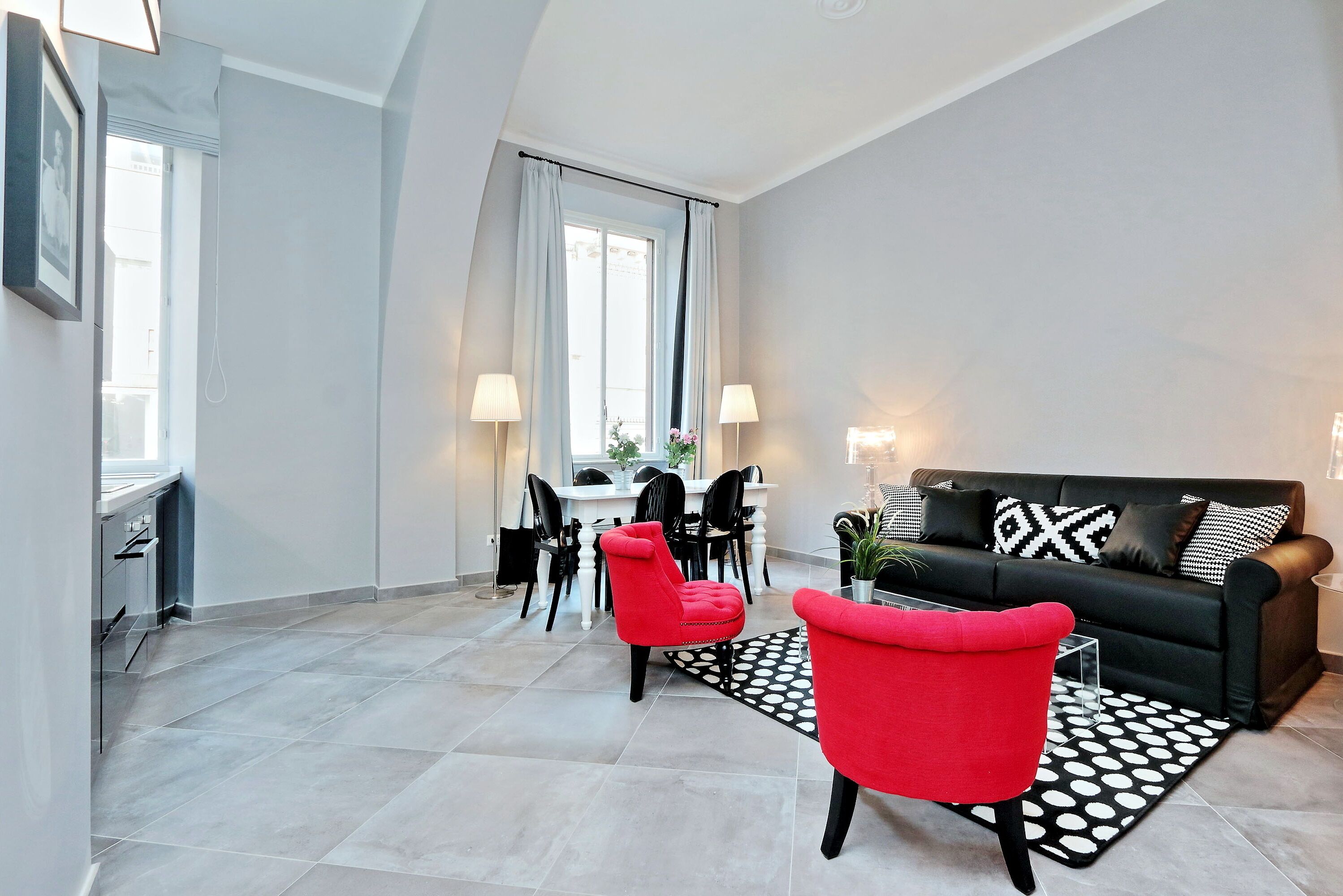 A modern living room with a black sofa, red chairs, a dining area with a table and chairs, and a large window with curtains.