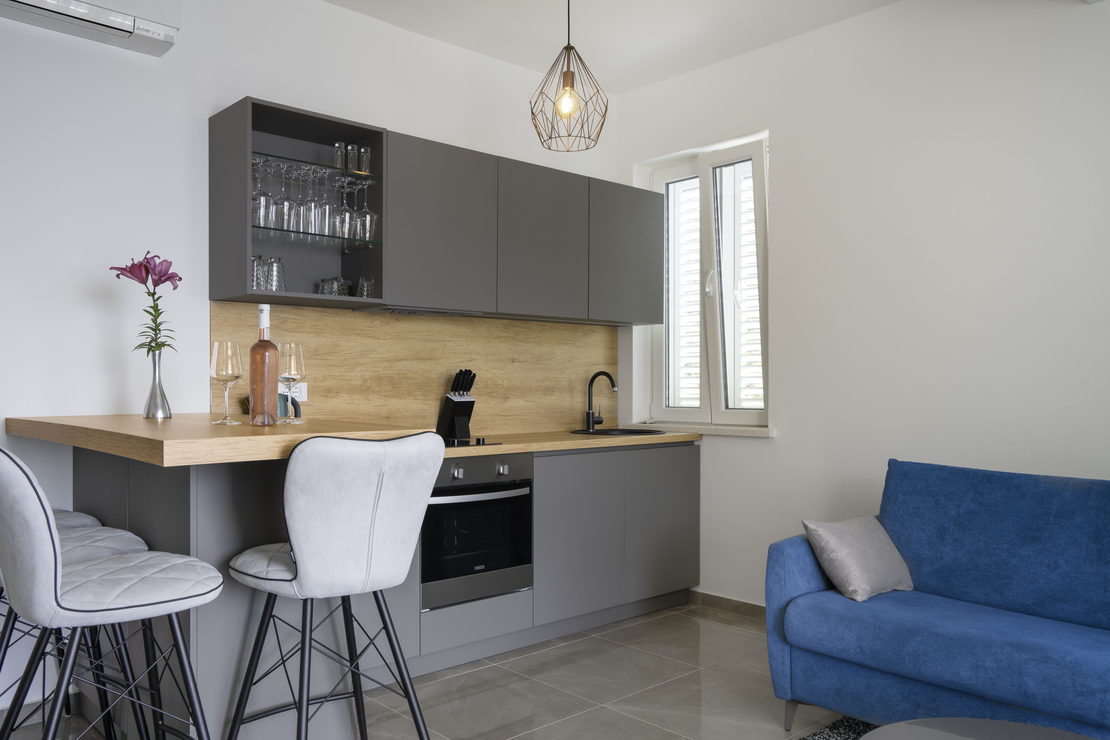A modern kitchen with grey cabinets, a wooden countertop, and a built-in oven. There is a blue sofa and a white chair in the adjacent living area.