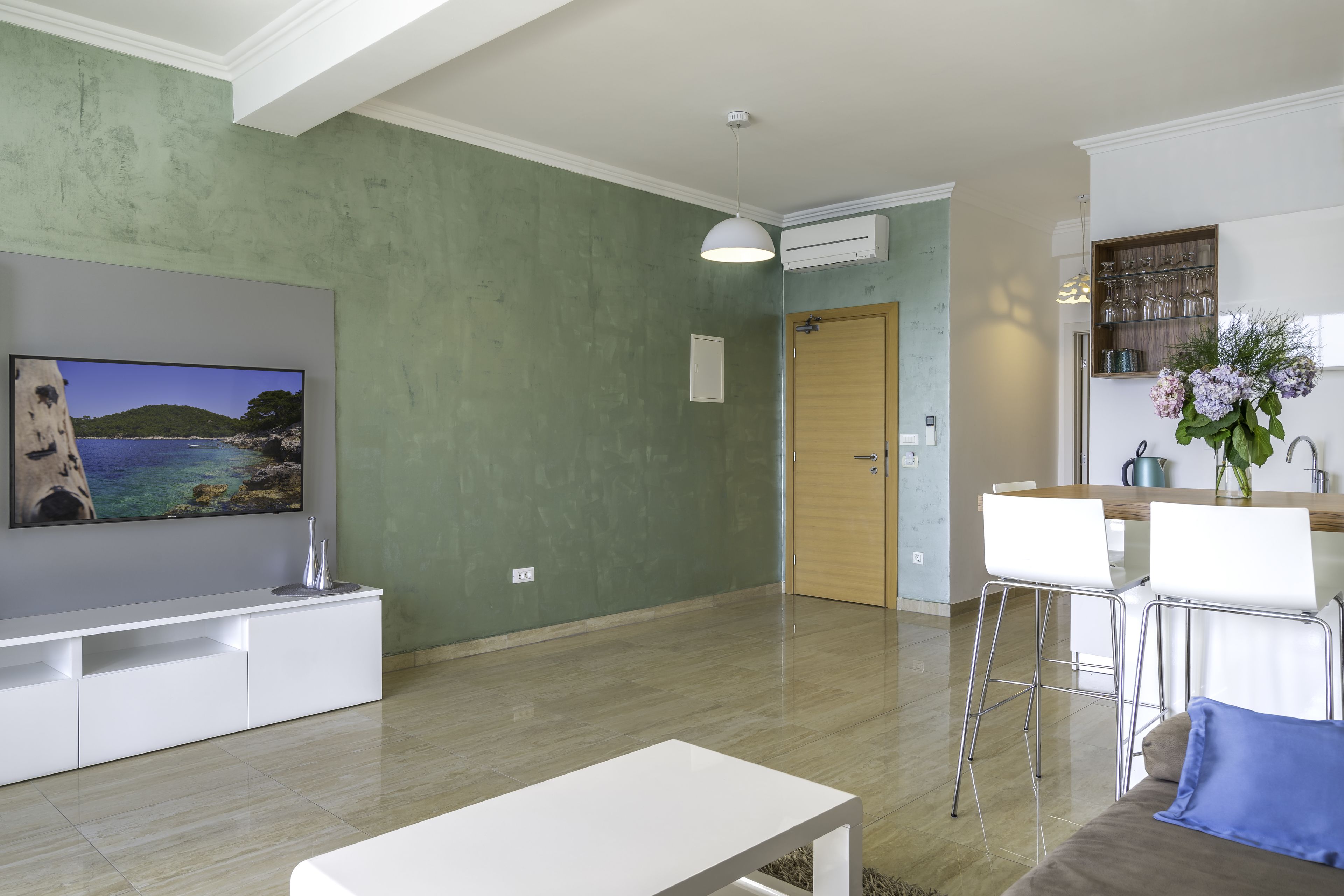 A modern living room with a flat-screen TV, a white entertainment unit, a dining area with white chairs, and a kitchen with a wooden countertop.