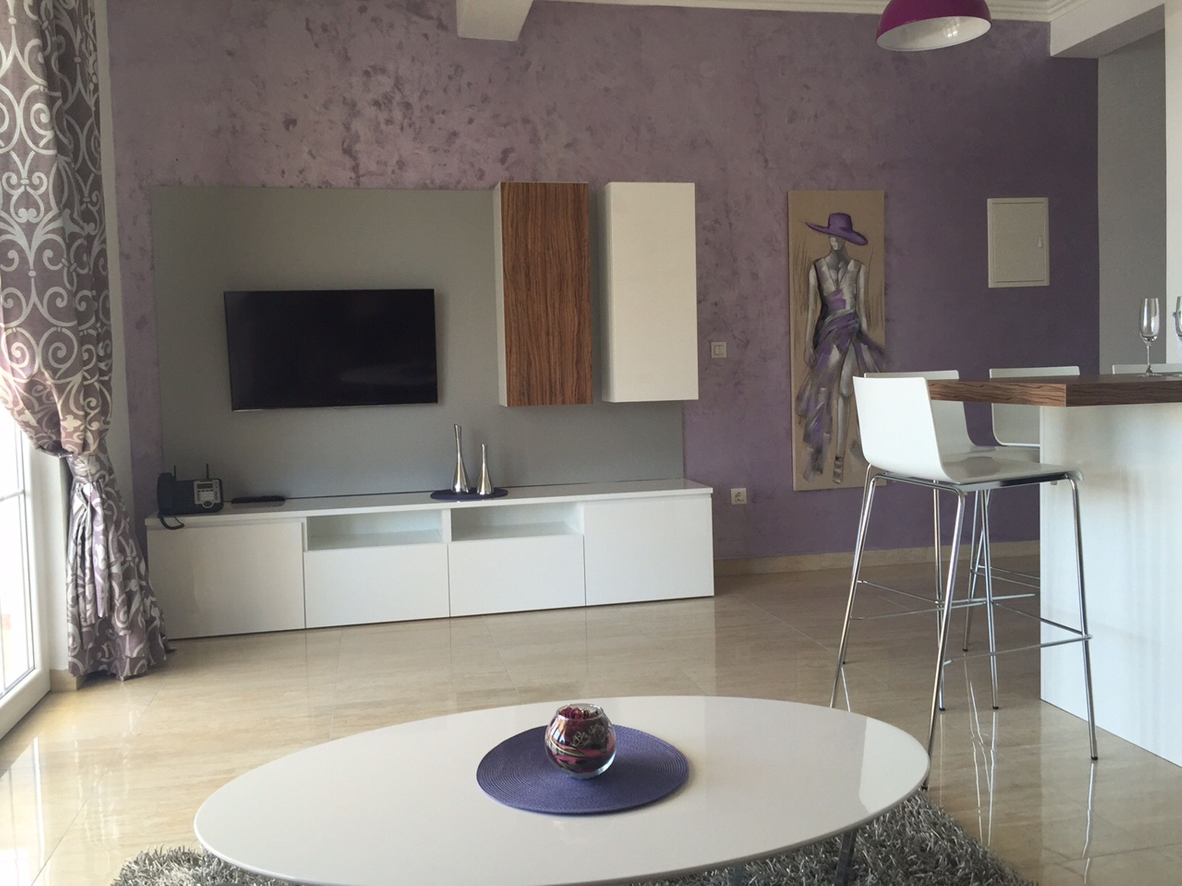 A modern living room with a round dining table, a flat-screen TV mounted on the wall, a kitchen area with a wooden countertop, and a painting on the wall.