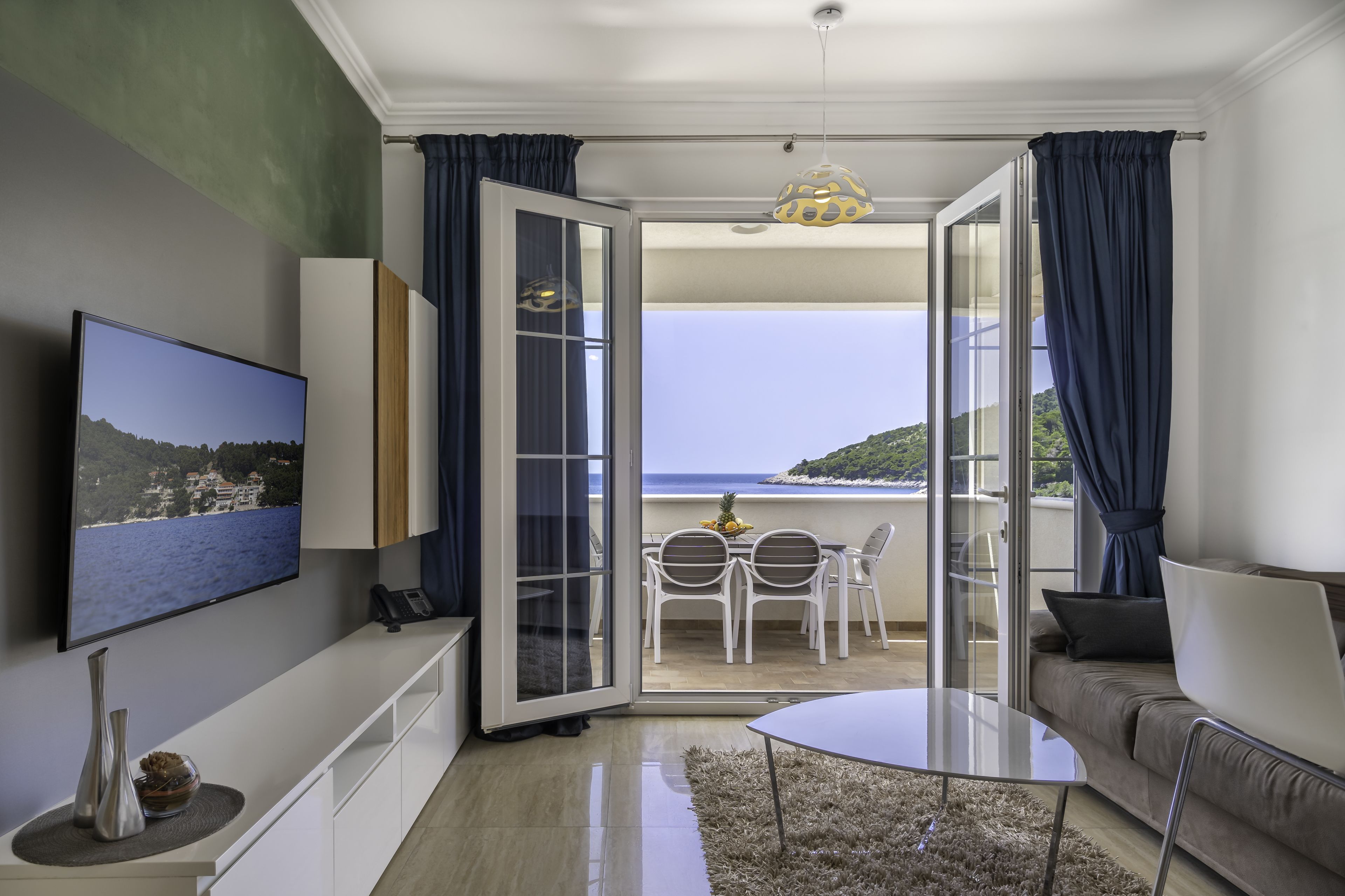 A modern living room with a TV, a sofa, a glass door leading to a balcony with a view of the sea.