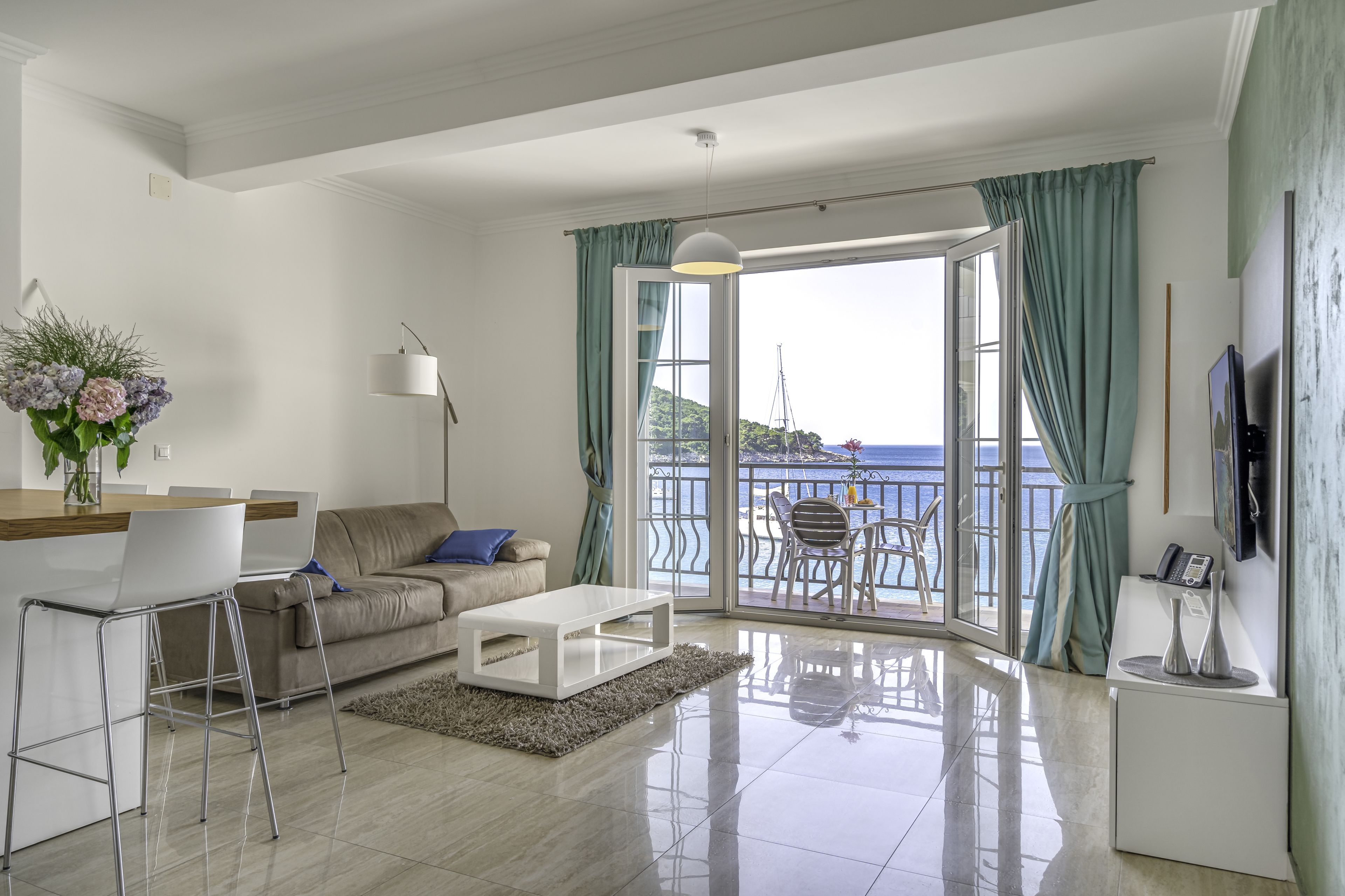 A modern living room with a sofa, coffee table, and bar area. A large window offers a view of the sea and a balcony with outdoor furniture.