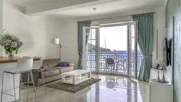 A modern living room with a sofa, coffee table, and bar area. A large window offers a view of the sea and a balcony with outdoor furniture.