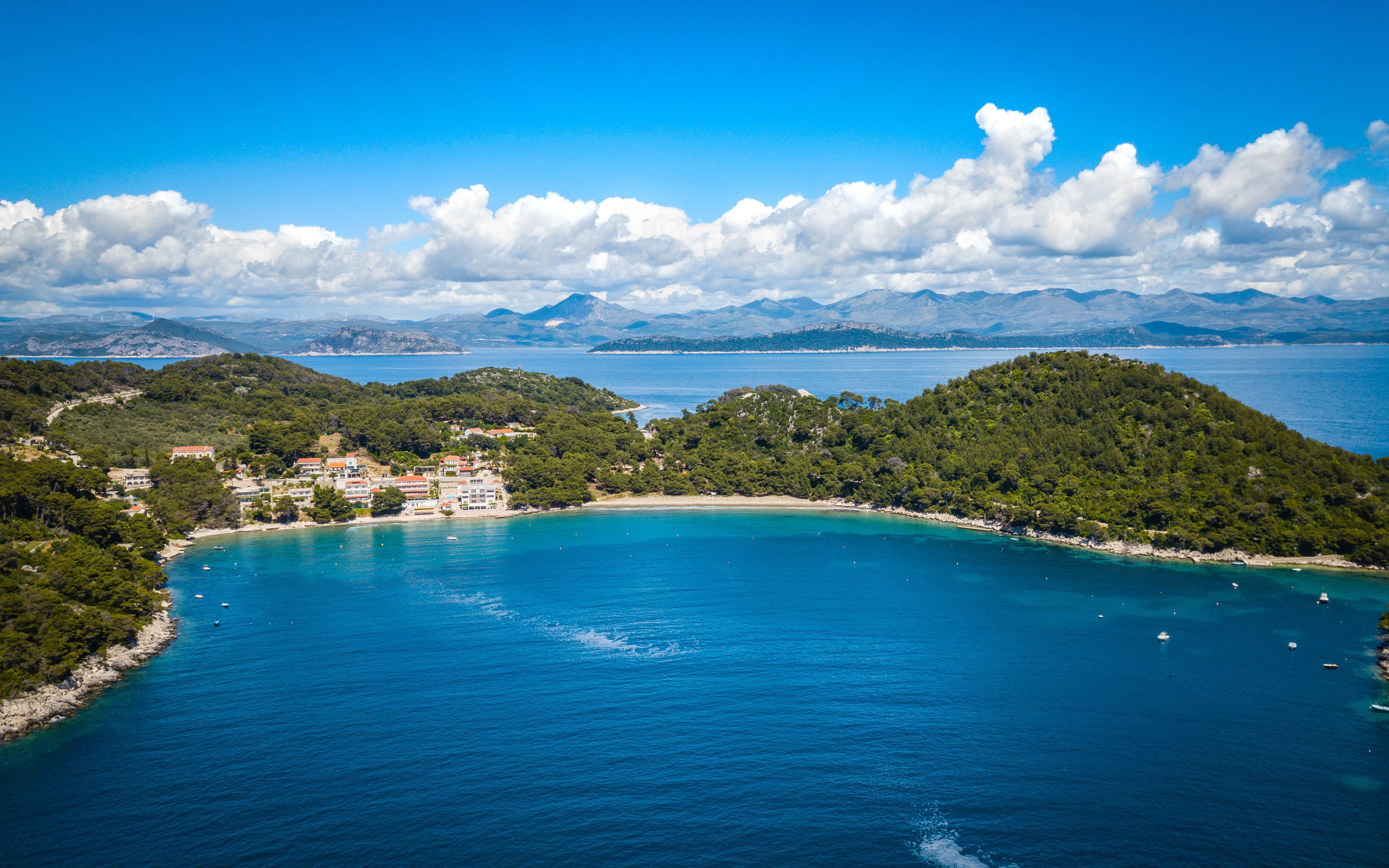 A coastal town with buildings along the shoreline, a clear blue sea, and mountains in the background.
