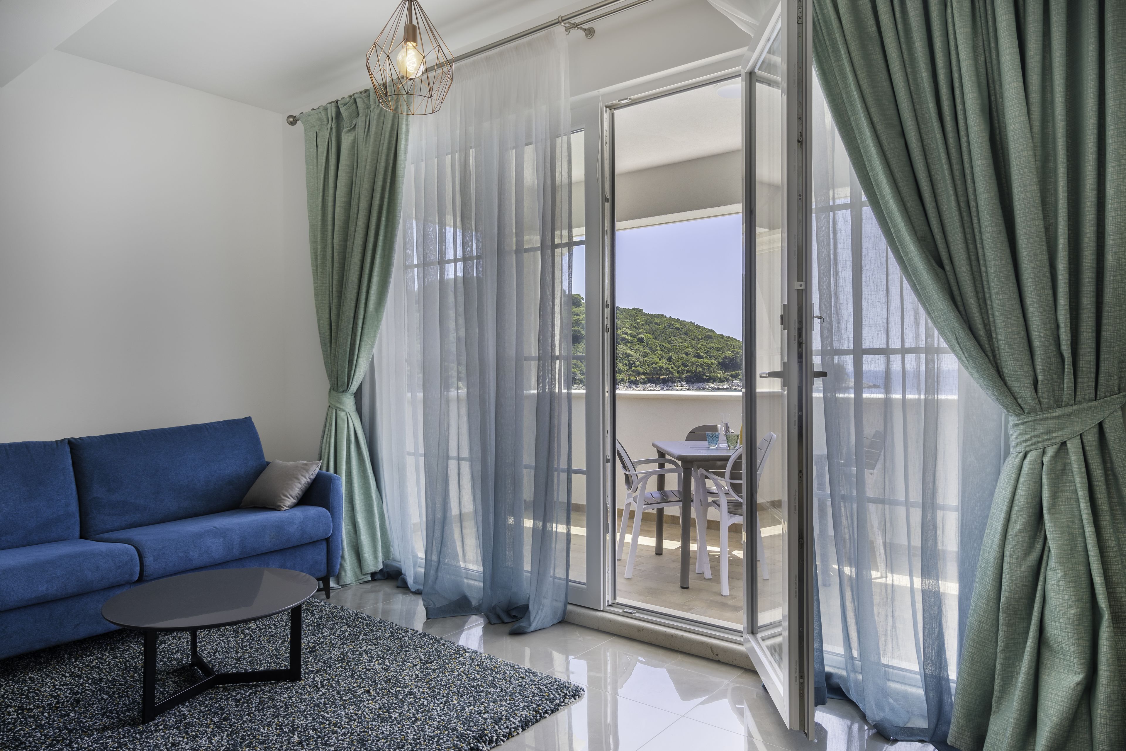 A room with a blue sofa, a small table, sheer curtains, and a view of a balcony with outdoor furniture.